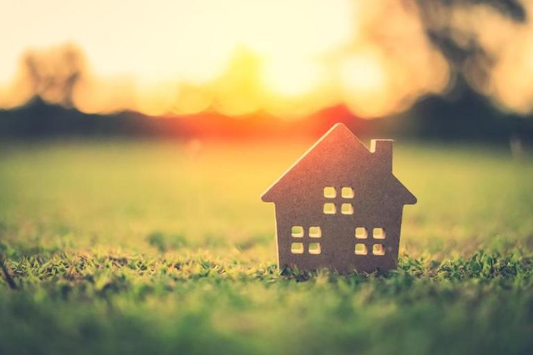 A model house sits on a grass lawn, with trees and hedges visible in the background. It's  sunset and the orange light shines through the windows of the house.