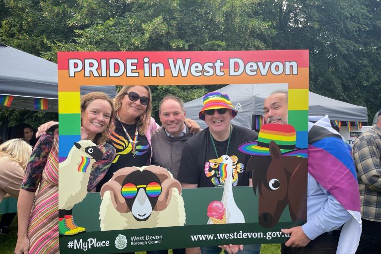 A group of people standing behind a selfie frame which reads PRIDE in West Devon