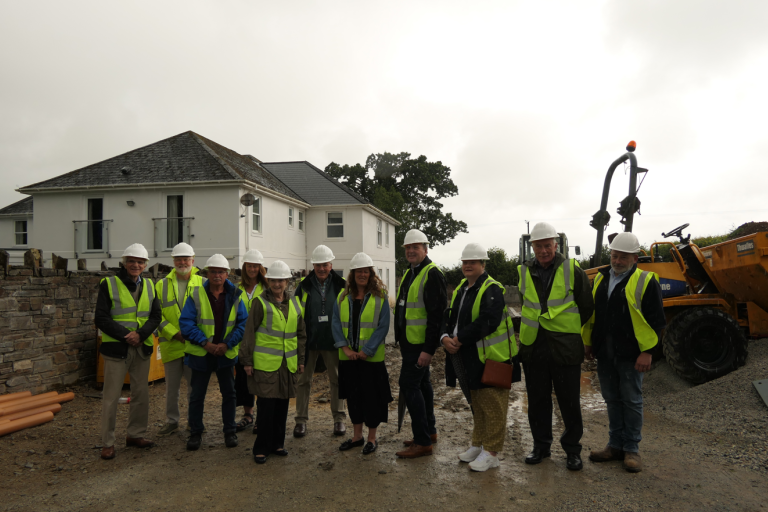 Group of eleven individuals in hard hats and high visibility vests standing in front of a house under construction, with a construction vehicle in the background.