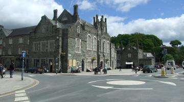 A road junction in Tavistock town centre, looking across a mini roundabout towards the Guildhall and the two square. The guildhall is a gothic style building built of Dartmoor granite. The sky is blue and partly cloudy.