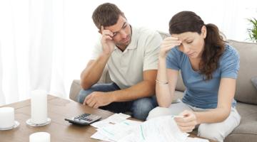 Man and woman sitting on sofa looking concerned as they look at bills piled on a wooden table