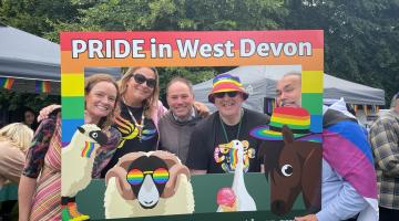 A group of people standing behind a selfie frame which reads PRIDE in West Devon
