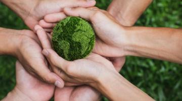 A group of hands clustered around a representation of the earth, made from grass and moss. The hands are carefully cradling the earth, as if they are caring for it together.