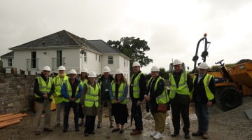 Group of eleven individuals in hard hats and high visibility vests standing in front of a house under construction, with a construction vehicle in the background.