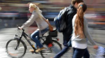 A blonde woman is riding a bicycle in a town centre, heading from right to left. A young couple, both with brown hair, walk in the opposite direction. They are deep in discussion.