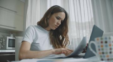 A young woman, sat at her kitchen table, looking through her energy bills. She has a concerned look on her face, as if the bills are more expensive than she imagined.