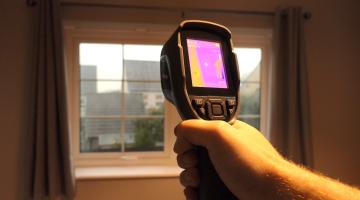 A man's hand holding a thermal imaging camera, pointed at window in a home. The camera is on and the display is giving an infrared readout. The window has plain grey curtains.