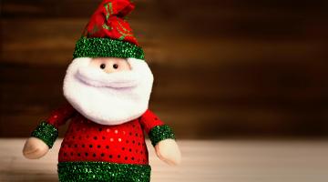 A picture of a crocheted Father Christmas toy, sat on a white table in front of a burgundy background.