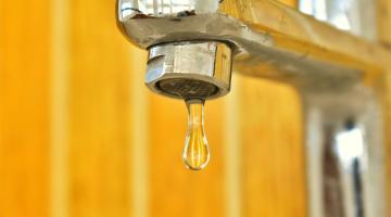 A dripping tap - the tap is silver in colour, and set against a blurred yellow background.